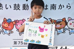 国体の横断幕の前で、白い体操服姿の男の子が手書きで「がんばれかごしま国体」の文字とイラストが描かれたスケッチブックを掲げ、ピースサインをしている写真