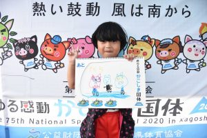 国体の横断幕の前で、小学生らしい児童が手書きのイラストが描かれたスケッチブックを掲げ、ピースサインをしている写真