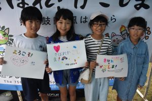 テントの下のブース前に、小学生くらいの児童4人が並んで立ち、それぞれが「きばれかごしま」などと書かれた応援ボードを持っている写真
