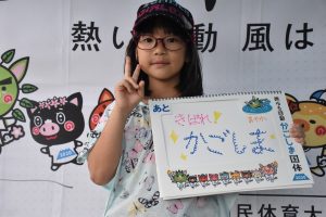 テントの下でメガネをかけ、キャップを被った黒髪の女の子が、右手でピースサインをして、カラフルな色で「きばれ！かごしま」と書かれたボードを持って立っている写真