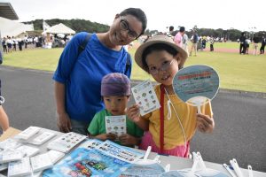 テントの前で、青いシャツを着た母親と、麦わら帽子を被りうちわを持った女の子、バンダナを巻いた男の子の三人家族が、ブースに並べられた商品越しに、笑顔でカメラに目線を向けている写真