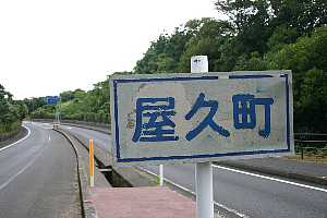 左側にカーブする舗装道路と緑豊かな木々が続く道路脇に「屋久町」と青文字で書かれた案内板が立っている写真