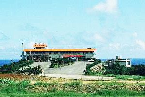 青空の下、海を背景に緩やかな坂道の先にオレンジ色の屋根を持つ2階建ての建物が立ち、周囲は緑の草地に囲まれた国民宿舎の外観写真