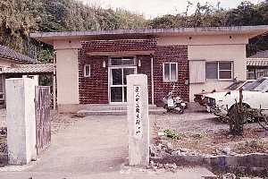 コンクリート造りの平屋の建物の前に、白い乗用車とスクーターが停まっており、建物の背後には鬱蒼とした緑の木々が生い茂る森が広がっている栗生診療所の外観写真