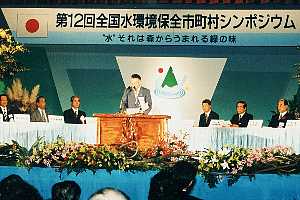 壇上中央に演台が置かれ、背後に「第12回全国水環境保全市町村シンポジウム」と書かれた横断幕が掲げられ、両側に複数の人物が並ぶ全国水会議の写真