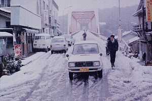 雪が積もった坂道に白い乗用車が走り、周囲には建物と停車中の車が並び、奥に赤い鉄橋が見える大雪の安房の写真