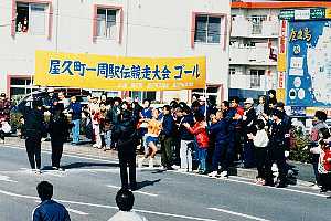 「屋久町一周駅伝競走大会 ゴール」と掲げられた黄色い横断幕の下、沿道に集まった大勢の観客が拍手や声援を送る中をオレンジ色のユニフォームのランナーがフィニッシュ地点へ駆け込んでくるカラー写真