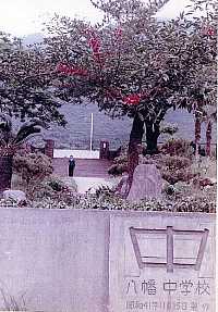 門柱に「八幡中学校」と刻まれた石碑があり、周囲には緑豊かな木々と赤い花をつけた木が立ち、奥に校門と広い通路が見える写真