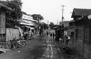 瓦屋根の木造家屋が並ぶ未舗装の通りを大人や子供が歩き、手前には自転車が停められ、奥には電柱や白い壁の建物も見える尾之間集落のモノクロ写真