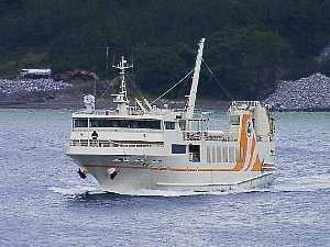 背景には緑の山と岩場が広がる沿岸に白とオレンジのラインが入ったフェリー太陽が穏やかな海面を航行している写真