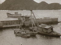 防波堤に複数の漁船が係留され、中央にはクレーンのような装置を備えた船があり、背景には山並みが広がる港の風景の写真