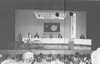 壇上に長机が並び、複数の人物が着席している会場で、背後には横断幕が掲げられた式典の写真