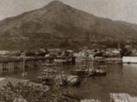 港の水面に複数の帆船が停泊し、背後には橋と建物が並び、そのさらに奥に大きな山がそびえる風景の写真