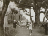 木々が並ぶ道を先頭のランナーがゴールテープを切り、後方に複数のランナーが続き、沿道では人々が旗や紙吹雪で応援している山岳競技会の写真