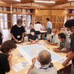 十数人の参加者が中央の大きなテーブルを囲み、広げられた地図や資料を見ながら、熱心にワークショップ形式で意見交換している写真