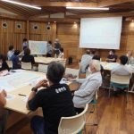 参加者の人々が資料を広げたテーブルに着席し、前方のスクリーン横では参加者のグループが立って、大きな地図を指しながら説明している様子の写真