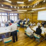 木造の梁と柱が露出した、高い天井の広い室内で、たくさんの参加者が、大きな白い紙を広げた机を囲んで座ったり立ったりしながらゲームを行っている様子の写真