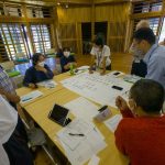 木製の壁に囲まれた室内で、複数の参加者が大きな机を囲み、広げられた模造紙に書かれた内容を確認したり、話し合いをしたりしているワークショップの様子の写真