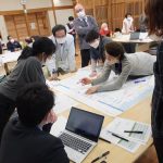 参加者たちが複数のテーブルに分かれて座り、手前のテーブルでは大きな用紙に一人の参加者が書き込もうとしている様子他のメンバーが身を乗り出してみているワークショップの様子の写真