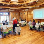 木造の天井と床の広い会場で、複数のグループに分かれてテーブルに座った参加者たちが、壁にかかった大きなスクリーンに映し出された資料に注目している写真