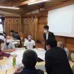 木造の室内で、複数のグループに分かれた参加者が、机を囲んで資料を広げ、活発に意見交換や作業を行っているワークショップの様子の写真