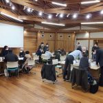 木目の壁と床、梁のある天井が特徴の円形の部屋で、約15人が小さなグループに分かれてテーブルにつき、熱心に議論や作業をしている様子を写した、ワークショップの写真