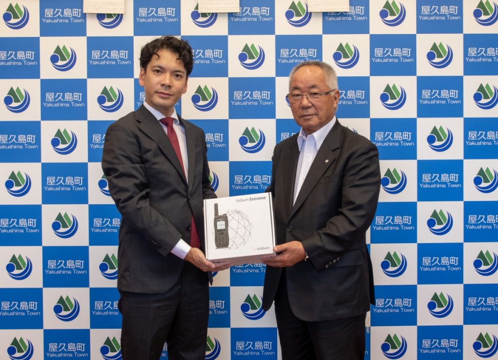 屋久島町のバックボード前で、スーツの男性と町長が並んで立ち、衛星電話のイラストが書かれた白い箱を両側から持っている記念写真