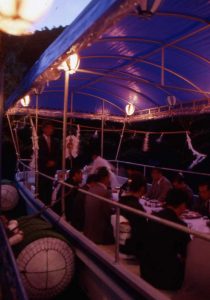 薄暗い屋外で船の青い屋根に付けられた複数のライトが灯り、船の上ではテーブルを囲んで座った人々が宴席を楽しんでいる様子の写真