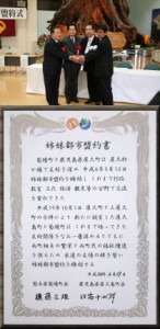 屋内の締結式会場で、背後に巨大な切り株のような木のオブジェとテーブルがある中で、スーツを着用した4人の男性が中央で握手を交わし記念撮影をしている写真と、その下に姉妹都市盟約書の本文と関係者の署名が記された書面が合成された、盟約締結の様子を伝える写真