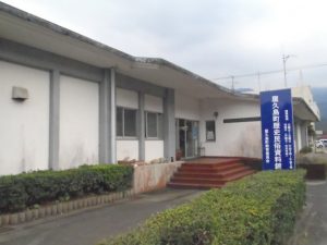 白壁の平屋建ての建物の正面には茶色の階段があり、その前には青い縦長の看板が立っている、生け垣で囲まれた屋久島町歴史民俗資料館の外観を捉えた写真