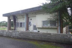 薄いクリーム色の平屋の建物が、コンクリート製の低い塀の奥に建っており、建物の左側には茶色い柱が複数並び庇を支えている住宅の写真