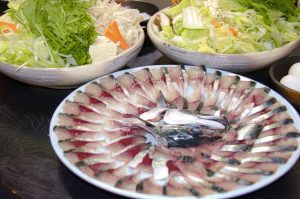 薄造りのサバの刺身が大皿に花のように盛られ、後ろにはたくさんの野菜や豆腐、きのこが盛られた大きな皿が2つ並んでいる写真