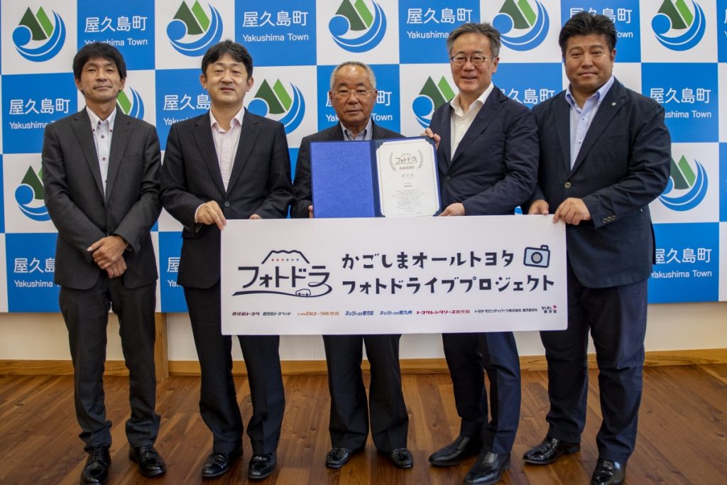 青と白の屋久島町のロゴが並ぶ背景の前で、スーツ姿の五人の男性が横一列に立ち、中央の町長が認定証が入った青い書類ケースを持ち、手前の「かごしまオールトヨタ フォトドラ フォトドライブプロジェクト」と書かれた白いボードと一緒に写した記念写真