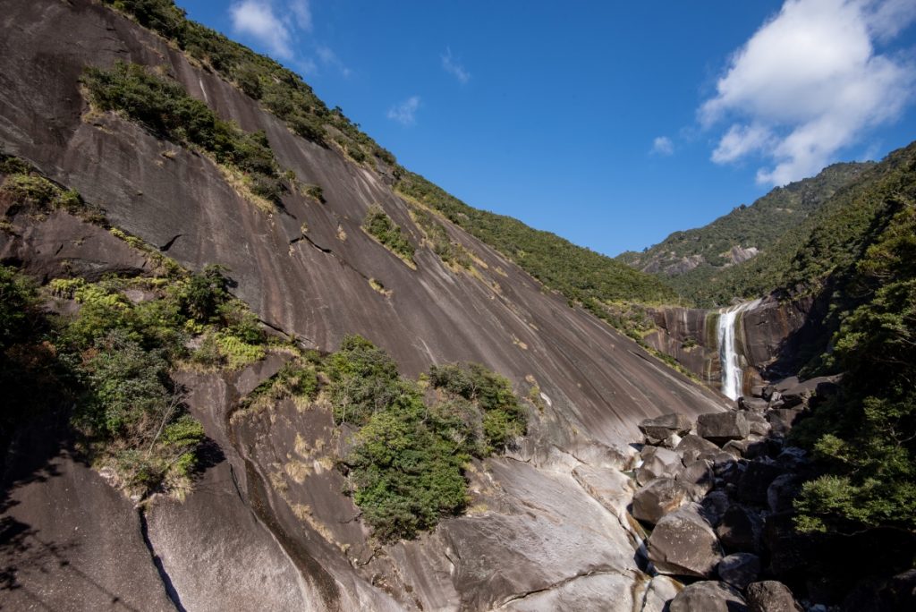 山間の灰色の岩肌の斜面の奥に、白い筋となって流れ落ちる滝と大小の岩がゴロゴロと転がる谷が見える雄大な自然風景の写真