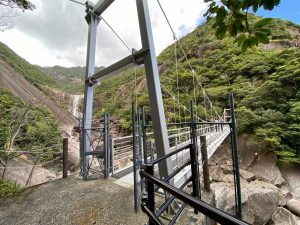 鋼鉄製の高い支柱とケーブルで構成された吊り橋が、森林に覆われた急峻な山々と岩がゴロゴロと転がる谷を渡って架けられており、奥の断崖には滝が流れ落ちている壮大な自然景観の写真