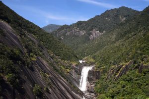 緑に覆われた山々に囲まれた深い谷の中を、白い水が幅広く傾斜した一枚岩の斜面を伝って勢いよく流れ落ちており、両側の山肌は黒っぽい岩が露出している壮大な滝の自然風景の写真