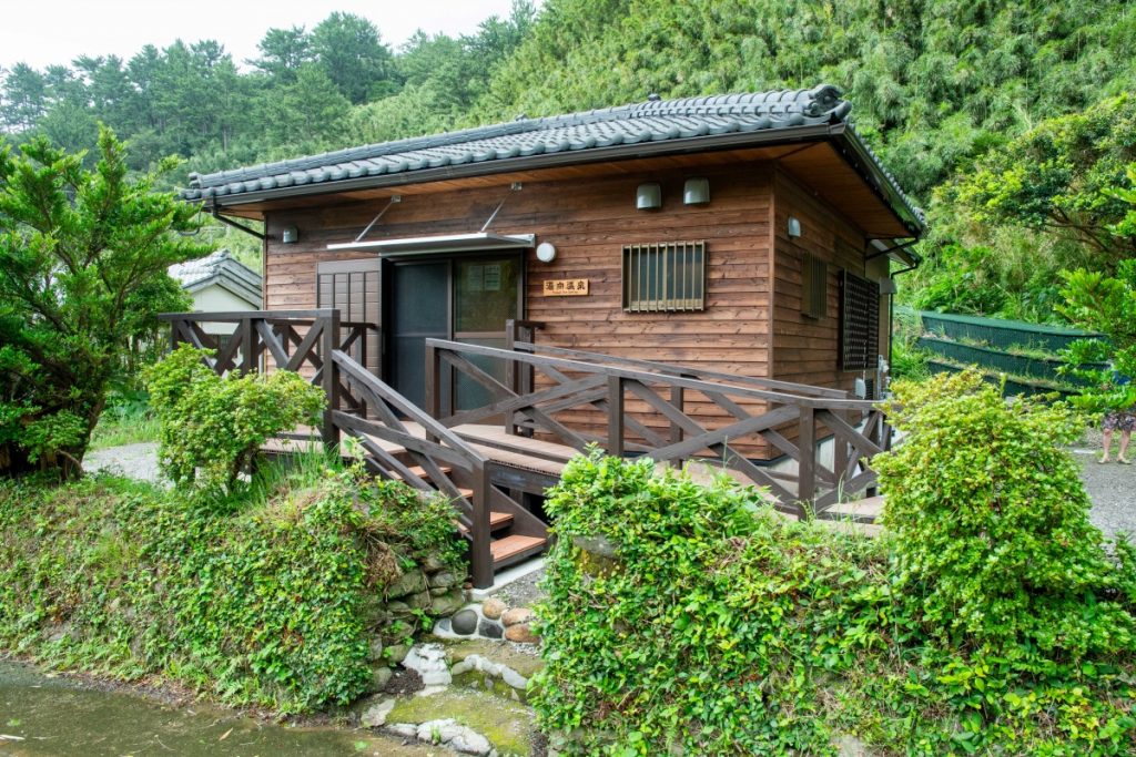 深い緑の木々に囲まれた、切妻屋根と焦げ茶色の板壁が特徴の、和風で趣のある木造平屋の建物を正面から捉えた写真