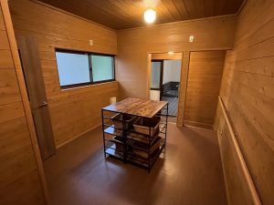 明るい木目の板壁と天井が特徴の室内で、部屋の中央に黒い金属フレームと木製の天板を備えた収納棚が配置され、その奥の開いた戸の向こうに湯船が見える脱衣所の写真