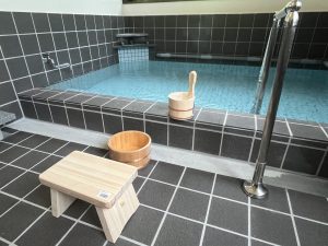 光沢のある黒いタイル張りの床と壁が特徴の浴室で、木製の風呂椅子と桶が置かれ、手前に手すり付きの銀色のポールがある、青いタイル張りの湯船が満たされた温泉の写真