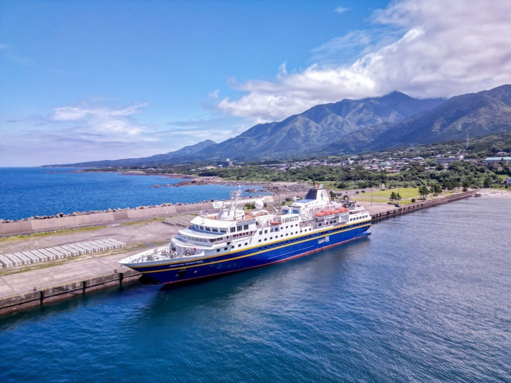 青い海に浮かぶ青と白を基調とした大型のクルーズ船が埠頭に停泊しており、その背景には家屋が立ち並ぶ港町と緑の山々がそびえ立つ雄大な風景を空撮した写真