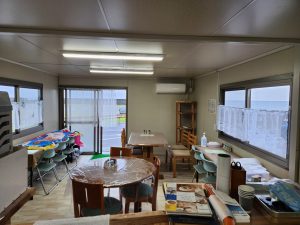 海辺に建つ簡素なプレハブ造りの建物内部を写したスナップ写真で、窓の外には灰色の空と海が広がり、室内にはテーブルや椅子、流し台などが配置された簡素な空間の写真
