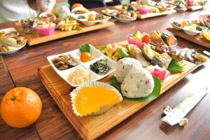 色とりどりのたくさんの料理が所狭しと並んだ木製のトレーや小皿、ミカンが長テーブルに複数並べてある写真