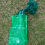 芝生の上に緑色の防水性ジッパーバッグが置かれ、袋の中央には白文字で「powerful & tight zip bag」と印字され、上部からは結ばれた濃緑色の廃棄物袋が出ている携帯トイレ廃棄セットの写真