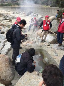 岩場にある温泉の源泉のような水溜まりから白い湯気が上がり、赤いベストを着たスタッフと数名の観光客が周りに集まっている様子の写真