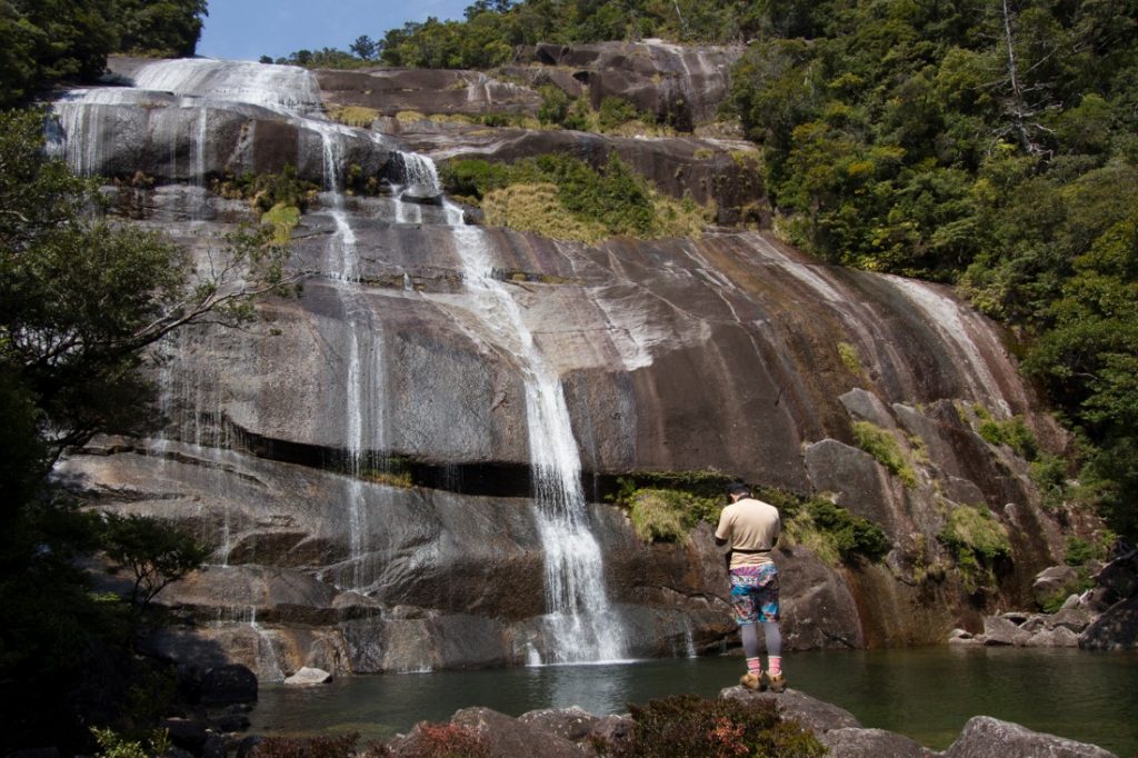 緑に囲まれた岩壁の岩肌を滑り落ちる複数の白い滝が、手前の水面に流れ込んでおり、対岸の岩の上に一人の人物が立っている写真