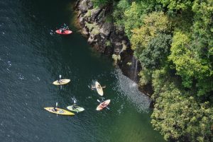切り立った岩肌から流れる小さな滝の近くで、6つのパドルボードなどが水面を漕ぎ進んでいる様子を、高い位置から俯瞰で捉えた写真
