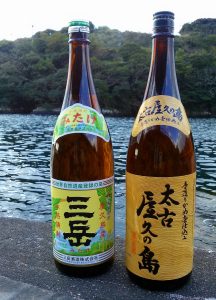 緑の森の前を流れる川を背景に、「三岳」「太古 屋久の島」のラベルが貼られた2本の茶色の酒瓶が並べて置かれている写真