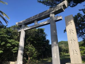 右側に「益救神社」の文字が刻まれた細い石碑があり、木々に囲まれて石造りの鳥居がそびえたっている写真