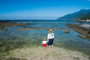 浅瀬の海岸で帽子をかぶった大人と子どもが手を繋ぎ海に足を入れている後ろ姿、透明な水と遠くの青い海、山並みが広がる風景の写真