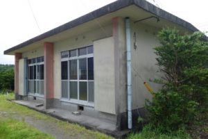 薄いクリーム色の平屋の建物で、正面にはアルミサッシの大きな窓が2枚あり、窓の左右にはピンクのアクセントが入った柱がある建物外観の写真